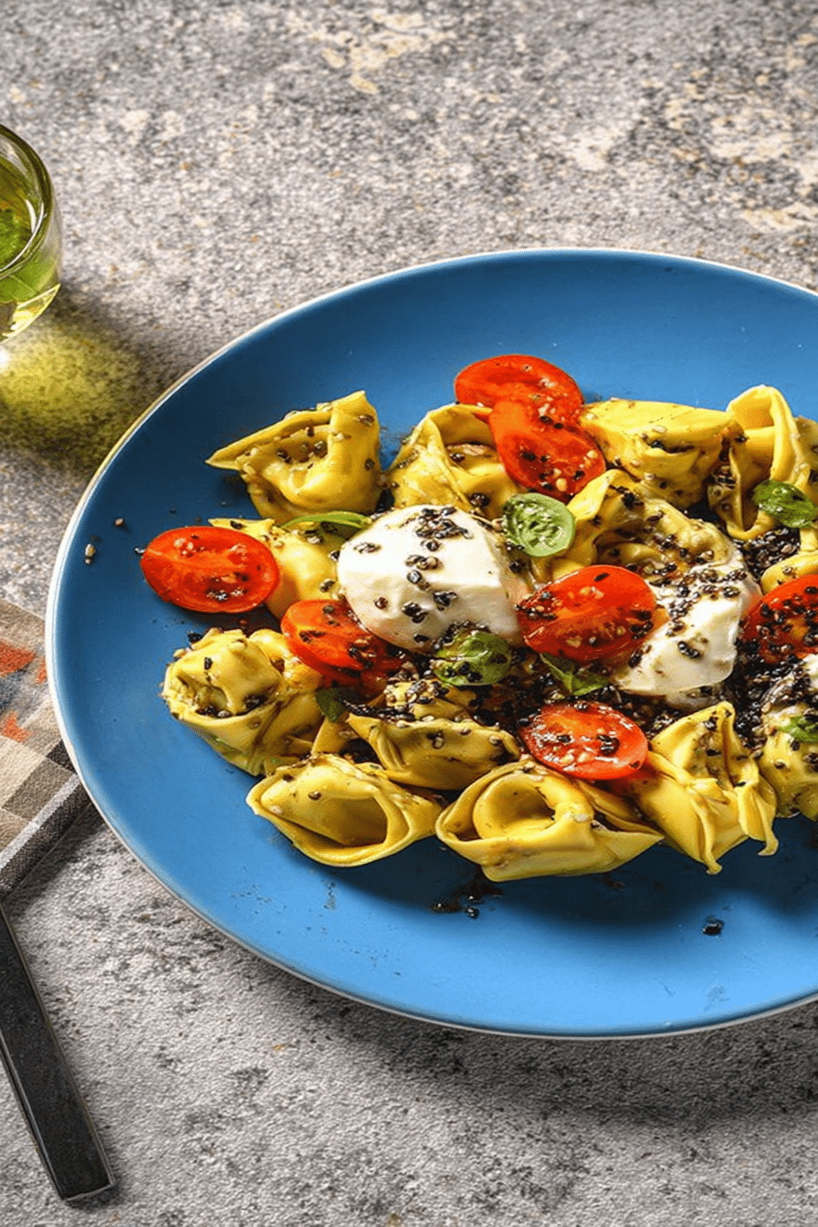 Frischer Tortellini-Salat mit Mozzarella, Cherry-Tomaten und Oliven-Pinienkern-Crunch auf einem Tisch.