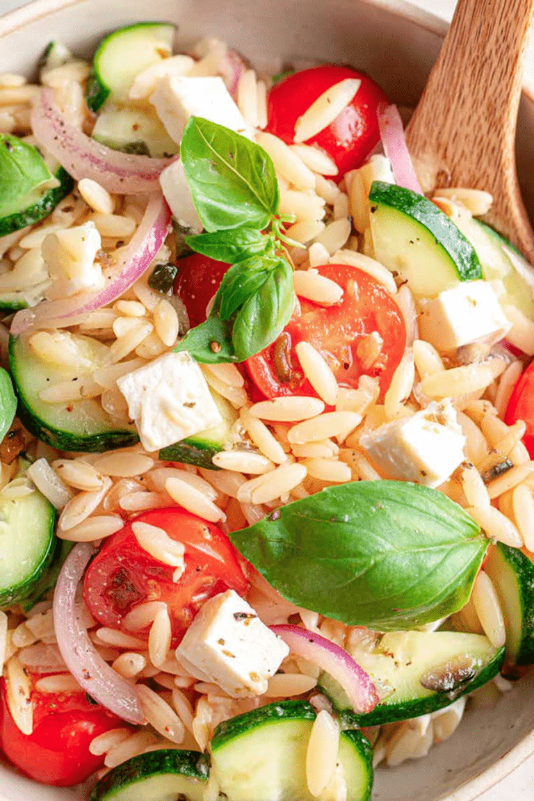 Ein bunter Kritharaki-Salat mit Feta und Gemüse auf einem Holztisch, dekoriert mit frischen Basilikumblättern, ideal für Sommergerichte.