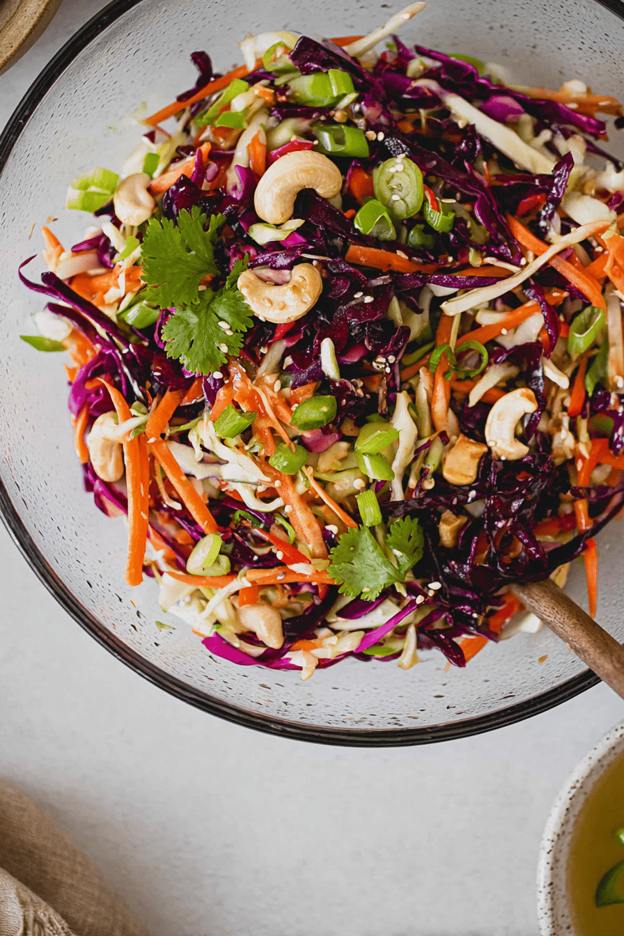Frisch zubereiteter asiatischer Krautsalat in einer bunten Schüssel auf dem Tisch, umgeben von frischen Zutaten wie Kohl, Karotten und Cashewnüssen.